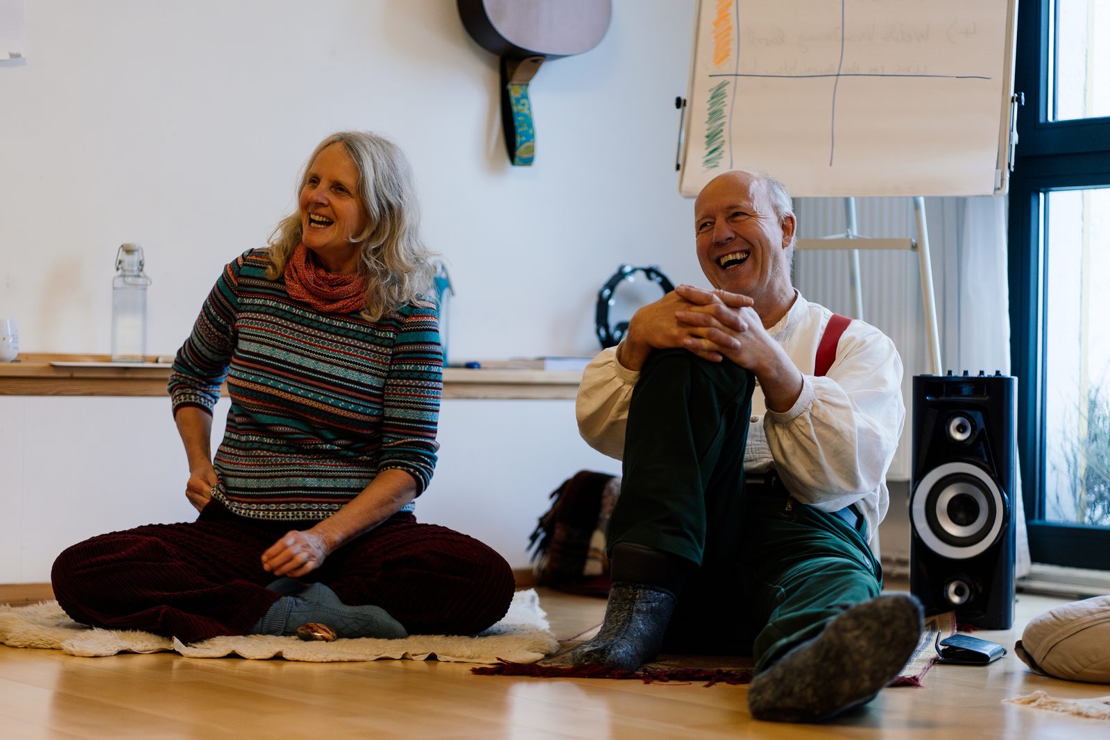 Uta Johanna Walle und Stephan Josef Dick sitzen lachend auf dem Boden eines Seminarraums